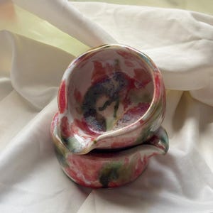 May include: Two stacked ceramic bowls featuring a glossy finish with a mottled pattern of pink, red, green, and dark blue hues. Each bowl has a small, integrated pouring spout on the rim, showcasing a rounded, organic shape and smooth texture.