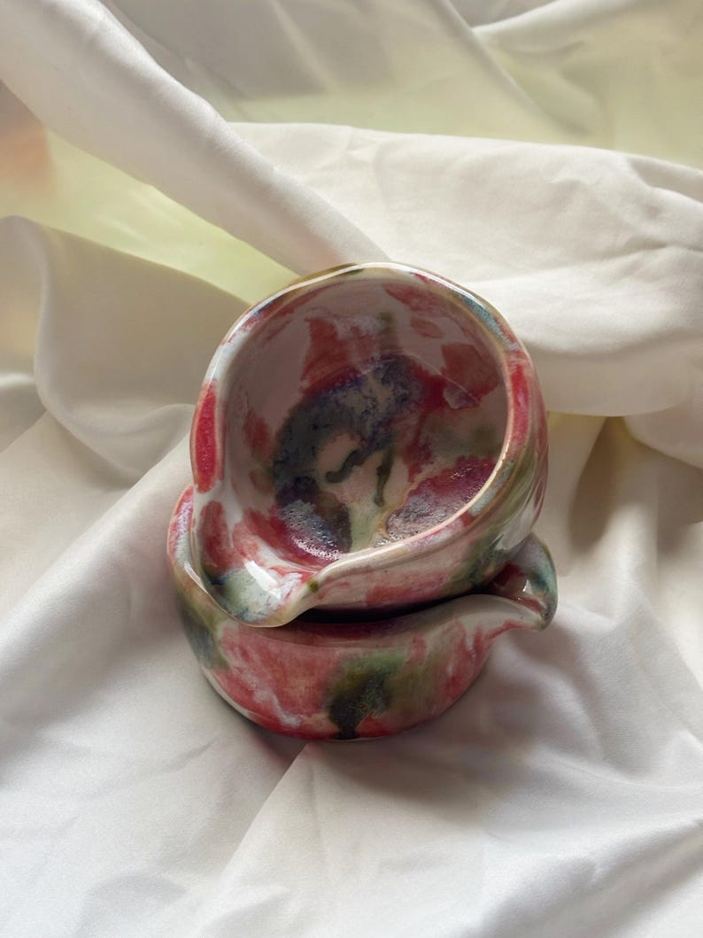 May include: Two stacked ceramic bowls featuring a glossy finish with a mottled pattern of pink, red, green, and dark blue hues. Each bowl has a small, integrated pouring spout on the rim, showcasing a rounded, organic shape and smooth texture.