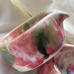 May include: A ceramic creamer pitcher featuring a glossy finish with a marbled pattern of blended pink, deep green, and white colors. The vessel has a curved rim with a small integrated pouring spout and a smooth, rounded body shape.