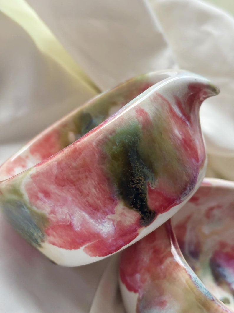 May include: A ceramic creamer pitcher featuring a glossy finish with a marbled pattern of blended pink, deep green, and white colors. The vessel has a curved rim with a small integrated pouring spout and a smooth, rounded body shape.