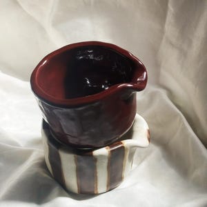 May include: Two stacked ceramic pitchers with irregular, hand-formed shapes. The top pitcher is a solid, glossy dark maroon. The bottom pitcher features a cream-colored base with vertical, hand-painted dark brown stripes and a small pouring spout.