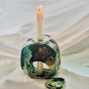 May include: A ceramic candle holder with a glossy, marbled glaze in shades of teal, brown, and black. The sculptural piece features a central opening and a top socket holding a lit cream-colored taper candle. A small, matching circular dish sits nearby.