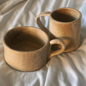 May include: Two beige ceramic mugs with speckled detailing. One mug is shorter and wider, while the other is taller and narrower. Both have curved handles and are set against a light blue fabric background. The mugs have a rustic, handmade appearance.