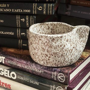 May include: A small, round, ceramic cup with a speckled brown and cream finish. The cup features a textured surface and a single, integrated handle on the side. It is positioned resting on top of a stack of hardcover books with visible text.