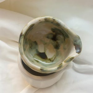 May include: A ceramic bowl with an irregular, organic rim and a glossy finish. The interior features a mottled glaze pattern in shades of sage green, cream, and dark brown, while the exterior base is a solid, smooth off-white ceramic.