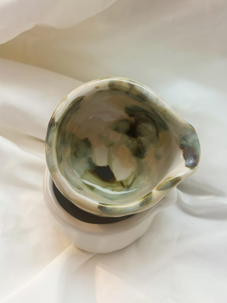 May include: A ceramic bowl with an irregular, organic rim and a glossy finish. The interior features a mottled glaze pattern in shades of sage green, cream, and dark brown, while the exterior base is a solid, smooth off-white ceramic.