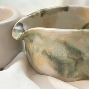 May include: A ceramic creamer pitcher featuring a hand-painted watercolor-style glaze in shades of sage green, olive, and soft cream. The vessel has a rounded body with a distinct pouring spout and a smooth, glossy finish throughout the exterior.