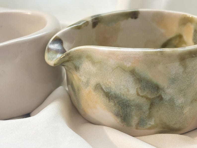 May include: A ceramic creamer pitcher featuring a hand-painted watercolor-style glaze in shades of sage green, olive, and soft cream. The vessel has a rounded body with a distinct pouring spout and a smooth, glossy finish throughout the exterior.