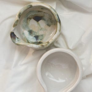 May include: Two ceramic bowls with pouring spouts. One bowl features a glossy white finish, while the other displays a marbled pattern of muted green, brown, and cream tones. Both vessels have a rounded shape and a smooth, reflective surface.