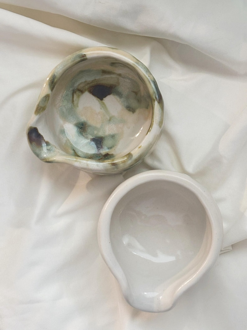 May include: Two ceramic bowls with pouring spouts. One bowl features a glossy white finish, while the other displays a marbled pattern of muted green, brown, and cream tones. Both vessels have a rounded shape and a smooth, reflective surface.