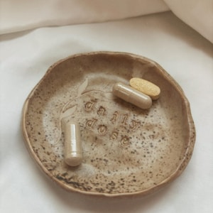 May include: A speckled, brown ceramic dish with the words "daily dose" embossed on the surface. The dish holds three capsules of varying colors. The dish has an organic, irregular shape and is likely handmade.