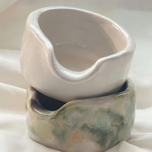 May include: Two stacked ceramic bowls with irregular, organic shapes and small pouring spouts. The top bowl is a solid off-white color, while the bottom bowl features a mottled, blended glaze of sage green, soft brown, and creamy white tones.
