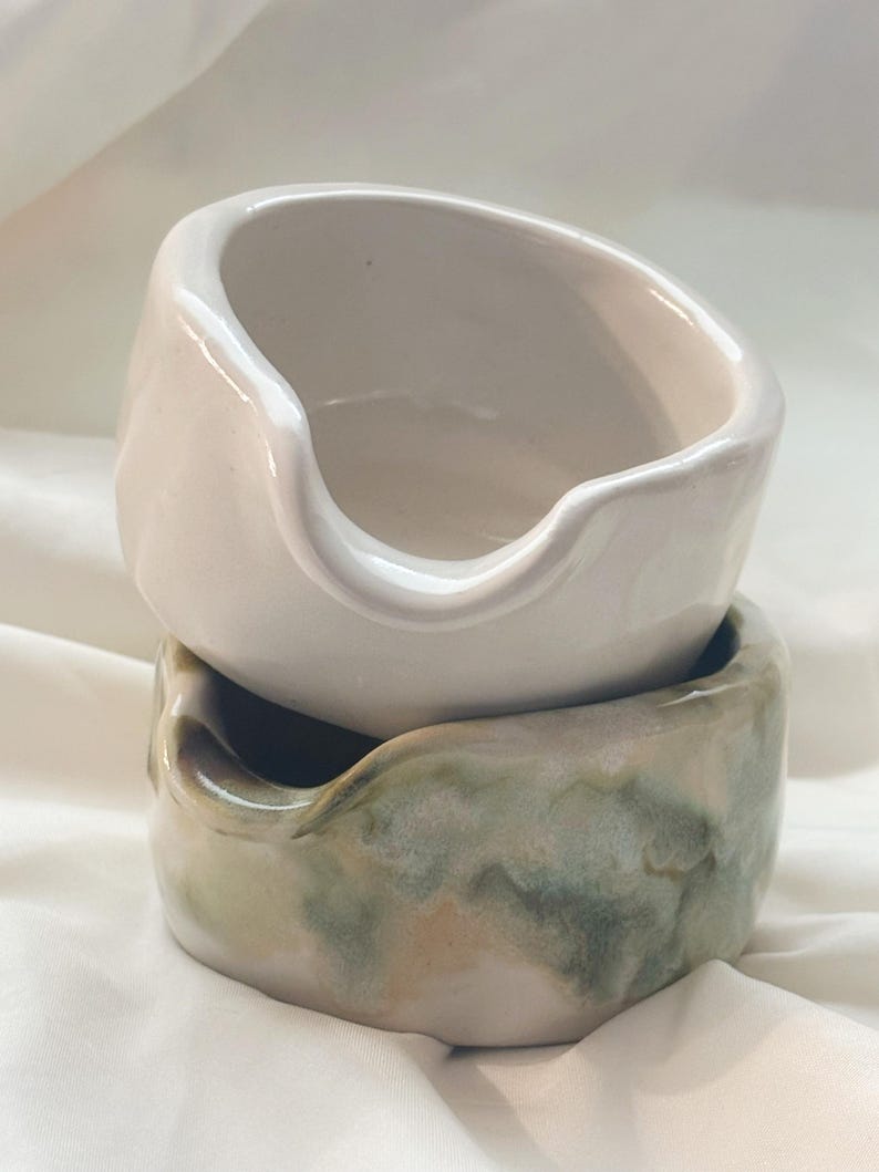 May include: Two stacked ceramic bowls with irregular, organic shapes and small pouring spouts. The top bowl is a solid off-white color, while the bottom bowl features a mottled, blended glaze of sage green, soft brown, and creamy white tones.