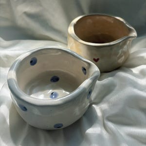 May include: Two small, hand-formed ceramic bowls with irregular, organic rims and pouring spouts. The front bowl is white with scattered blue dots, while the back bowl is a warm, glossy cream color featuring a single small red heart.