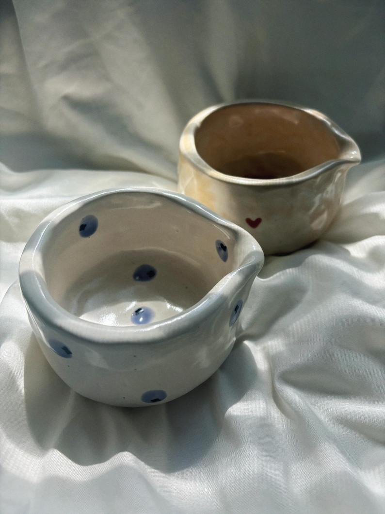 May include: Two small, hand-formed ceramic bowls with irregular, organic rims and pouring spouts. The front bowl is white with scattered blue dots, while the back bowl is a warm, glossy cream color featuring a single small red heart.