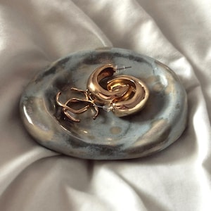May include: A small, oval-shaped ceramic dish with a glossy, mottled grey and blue glaze. Resting inside the dish are two gold-toned hoop earrings with a thick, rounded profile and a pair of delicate, thin gold-toned wire hoop earrings.