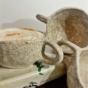 May include: Three handmade ceramic mugs with a speckled cream and brown finish. One mug is filled with a latte topped with cinnamon. The mugs feature rounded handles and irregular, organic shapes. They are stacked on top of two hardcover books.