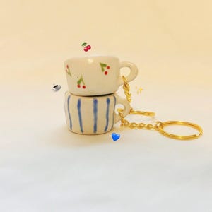 May include: Two ceramic teacup keychains stacked on top of each other. The top cup is white with red cherry accents. The bottom cup is white with blue vertical stripes. Both have gold keychains.