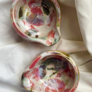 May include: Two small, round ceramic bowls with a glossy finish, featuring an abstract, marbled pattern in shades of pink, green, and white. Each bowl has a distinct, curved pouring spout on one side, creating an irregular, organic shape.