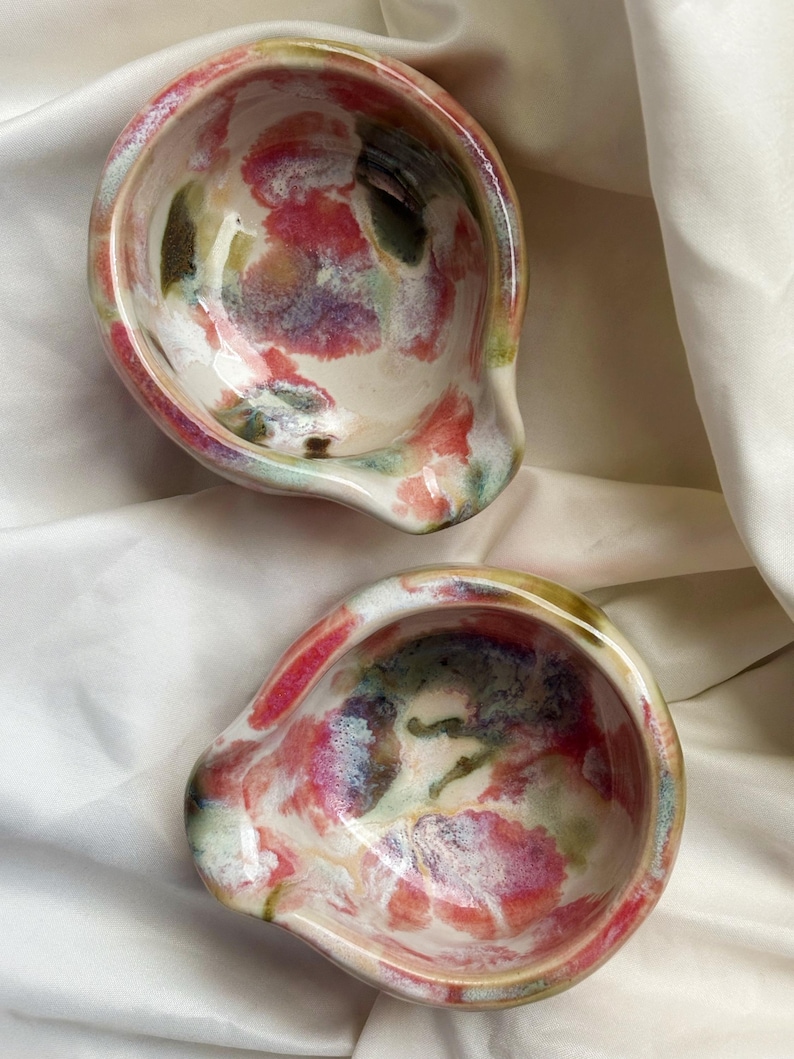 May include: Two small, round ceramic bowls with a glossy finish, featuring an abstract, marbled pattern in shades of pink, green, and white. Each bowl has a distinct, curved pouring spout on one side, creating an irregular, organic shape.