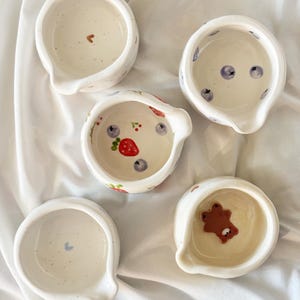 May include: Five small, white ceramic bowls with pouring spouts. Each bowl features unique hand-painted details inside, including a brown bear, a red strawberry with blueberries, a small brown heart, a blue heart, and scattered blue berries.