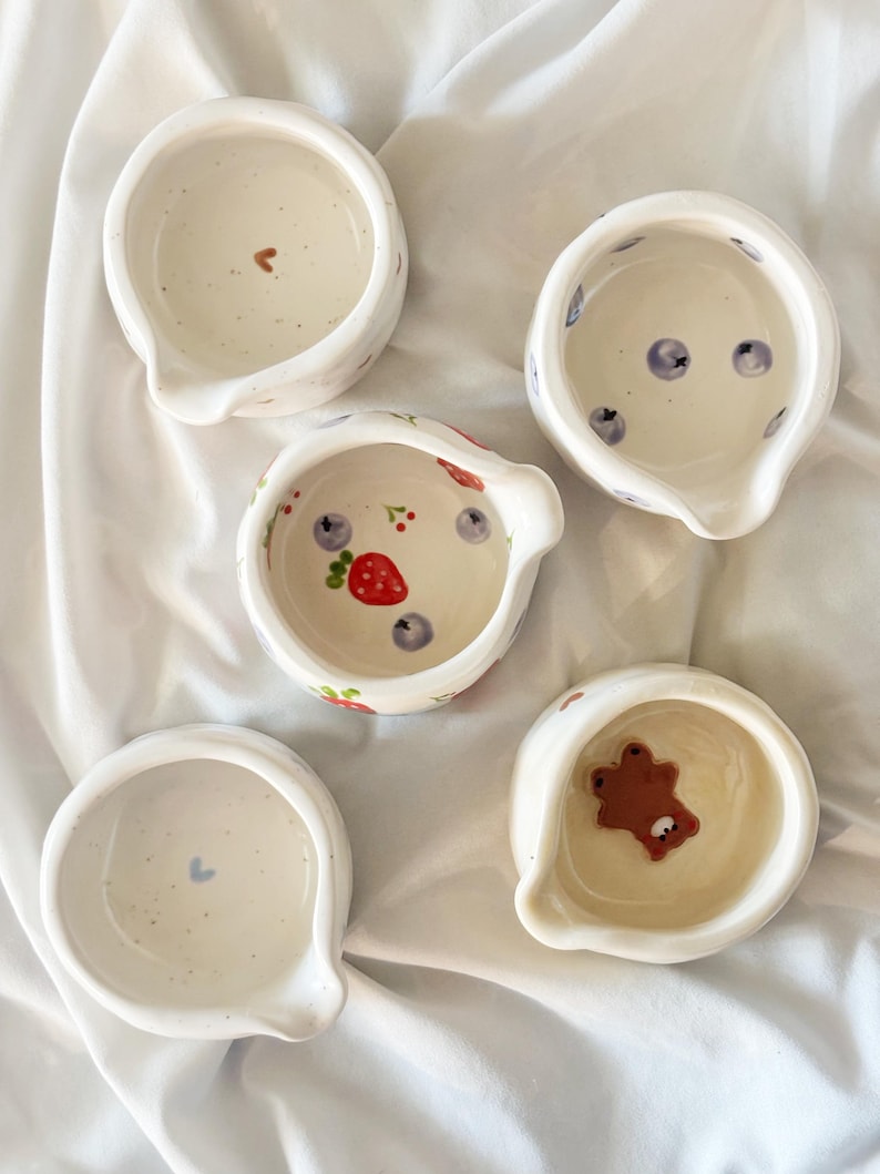 May include: Five small, white ceramic bowls with pouring spouts. Each bowl features unique hand-painted details inside, including a brown bear, a red strawberry with blueberries, a small brown heart, a blue heart, and scattered blue berries.