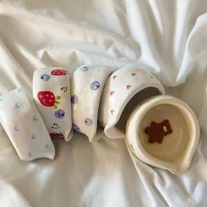 May include: A collection of five small, irregular ceramic bowls with various hand-painted designs. The patterns include blue hearts, red strawberries with blueberries, blue berries, brown hearts, and a single brown bear inside the final bowl.