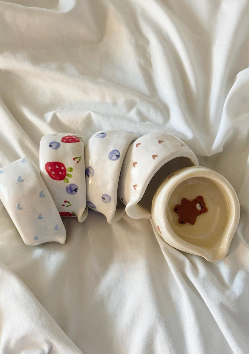 May include: A collection of five small, irregular ceramic bowls with various hand-painted designs. The patterns include blue hearts, red strawberries with blueberries, blue berries, brown hearts, and a single brown bear inside the final bowl.
