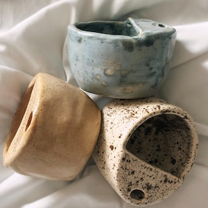 May include: Three ceramic wall planters in shades of blue, tan, and speckled beige. Each planter has a unique shape and a hole for hanging. The planters are arranged on a white fabric background.