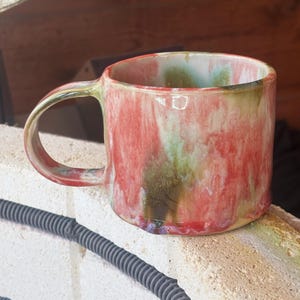 May include: A ceramic mug featuring a glossy, multi-colored glaze with swirling patterns of vibrant red, soft green, and creamy white. The mug has a rounded body, a smooth curved handle, and a wide opening, showcasing a fluid, drip-like finish.
