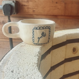 May include: A cream-colored ceramic mug with a rounded handle and a hand-drawn dark blue flower illustration enclosed in a scalloped square border on the front. The mug has a smooth, matte finish and a slightly irregular, organic shape.