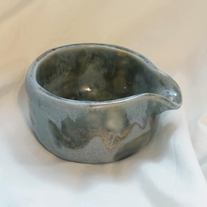 May include: A small, round ceramic pouring bowl featuring a mottled glaze in shades of slate blue, sage green, and earthy brown. The vessel has a smooth, curved rim with a distinct, integrated spout on one side for controlled pouring of liquids.