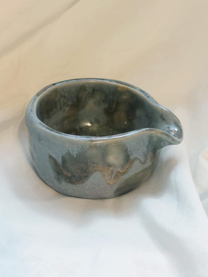May include: A small, round ceramic pouring bowl featuring a mottled glaze in shades of slate blue, sage green, and earthy brown. The vessel has a smooth, curved rim with a distinct, integrated spout on one side for controlled pouring of liquids.