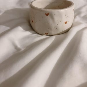 May include: A small, round, off-white ceramic bowl with an irregular, hand-formed texture. The exterior surface features a speckled pattern and small, hand-painted brown hearts distributed evenly around the circumference of the vessel.