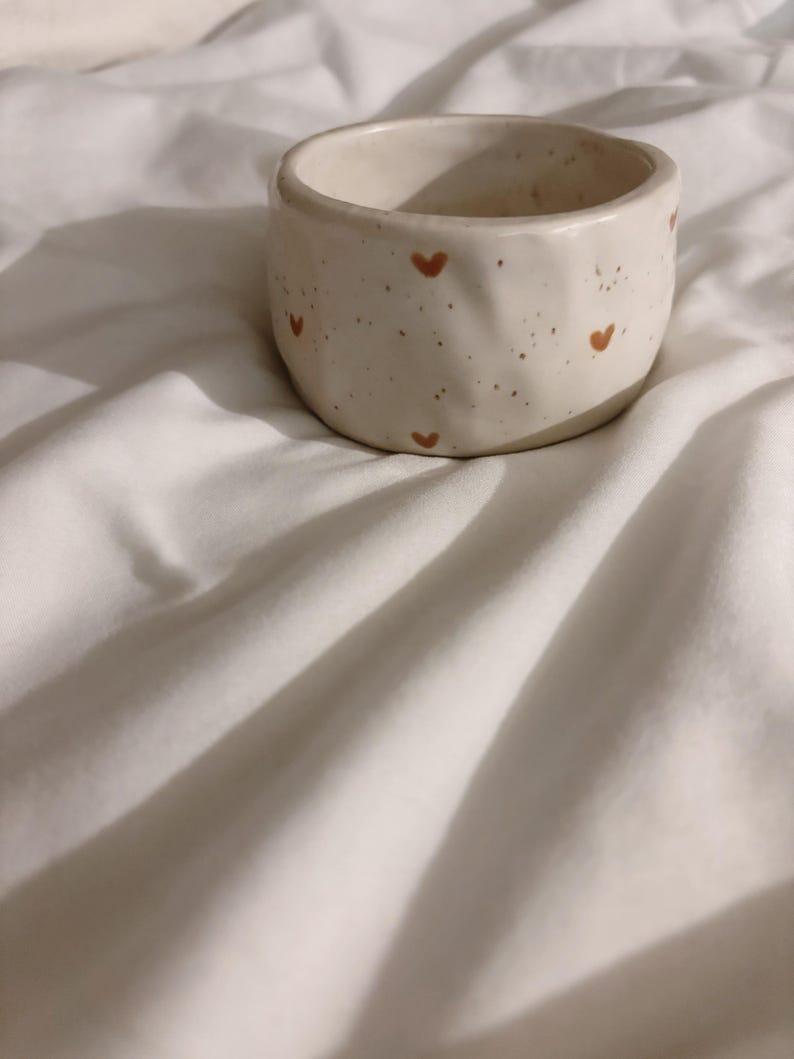 May include: A small, round, off-white ceramic bowl with an irregular, hand-formed texture. The exterior surface features a speckled pattern and small, hand-painted brown hearts distributed evenly around the circumference of the vessel.