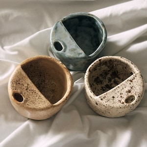 May include: Three ceramic bowls with a circular shape and a diagonal cut-out. The bowls are in shades of beige, speckled white, and blue-green. Each bowl has a small hole on the side. The bowls are placed on a white, textured surface.