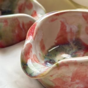May include: Two small, hand-formed ceramic bowls with an irregular, wavy rim. The bowls feature a glossy, white base glaze decorated with abstract, blended washes of coral pink, sage green, and deep blue pigments across the interior and exterior.