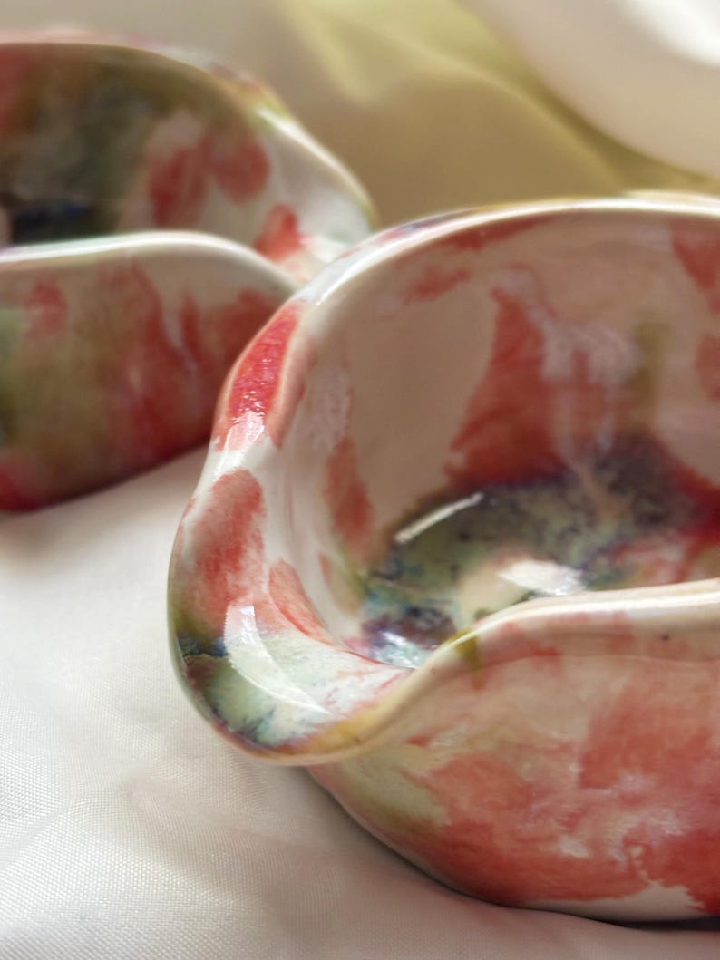 May include: Two small, hand-formed ceramic bowls with an irregular, wavy rim. The bowls feature a glossy, white base glaze decorated with abstract, blended washes of coral pink, sage green, and deep blue pigments across the interior and exterior.