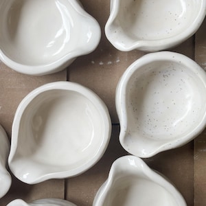 May include: A collection of small, white ceramic bowls arranged in a group. Each bowl features a rounded shape with a distinct pouring spout on one side. Some of the bowls have a smooth, solid white finish, while others display a speckled texture.