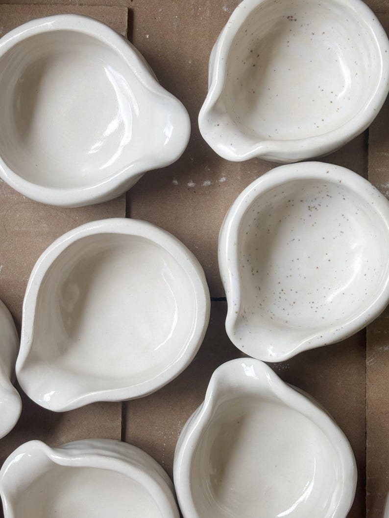 May include: A collection of small, white ceramic bowls arranged in a group. Each bowl features a rounded shape with a distinct pouring spout on one side. Some of the bowls have a smooth, solid white finish, while others display a speckled texture.