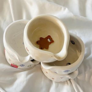 May include: A stack of three small ceramic bowls with pouring spouts. The top bowl features a brown bear figure at the bottom. The exterior of the bowls displays painted patterns including red strawberries, blue blueberries, and small brown hearts.