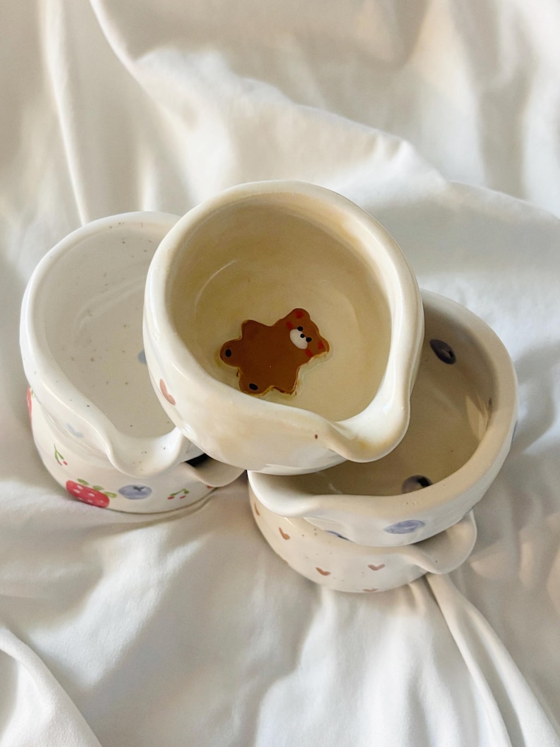 May include: A stack of three small ceramic bowls with pouring spouts. The top bowl features a brown bear figure at the bottom. The exterior of the bowls displays painted patterns including red strawberries, blue blueberries, and small brown hearts.