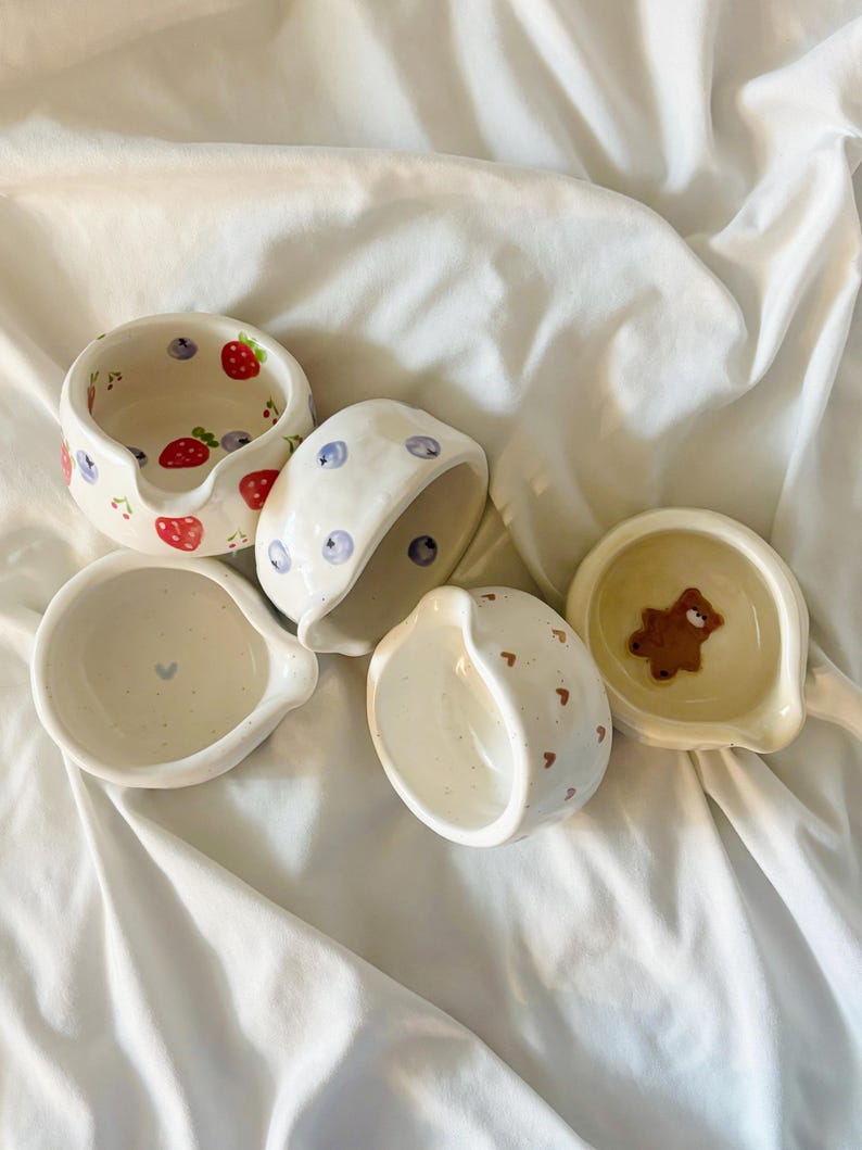 May include: Five small, round, white ceramic bowls with pouring spouts. One bowl features red strawberries and blue blueberries, one has blue blueberries, one has a small blue heart, one has brown hearts, and one contains a small brown bear figure.