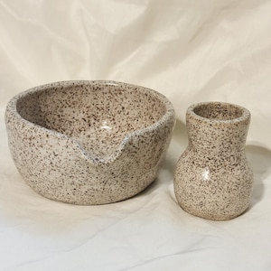 May include: A ceramic bowl with a pouring spout and a matching small vase, both featuring a speckled cream and brown finish. The bowl has a rounded shape with a distinct lip for pouring, while the vase has a narrow neck and a wider base.