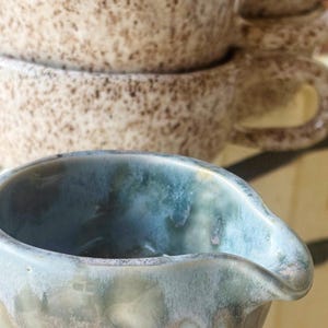 May include: A small ceramic creamer pitcher with a glossy blue and green reactive glaze featuring a pouring spout, positioned in front of a stack of three speckled beige ceramic mugs with handles and a textured, speckled finish.