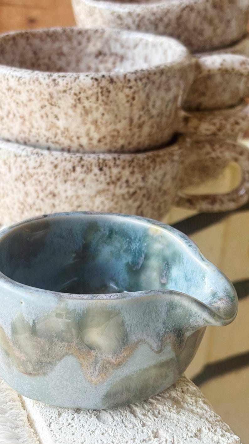 May include: A small ceramic creamer pitcher with a glossy blue and green reactive glaze featuring a pouring spout, positioned in front of a stack of three speckled beige ceramic mugs with handles and a textured, speckled finish.