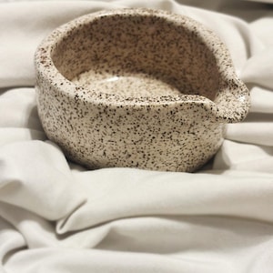 May include: A small, round ceramic bowl with a speckled beige and brown finish. The bowl features a distinct pouring spout on one side, a wide opening, and a smooth, curved exterior surface with a consistent pattern of dark brown flecks throughout.