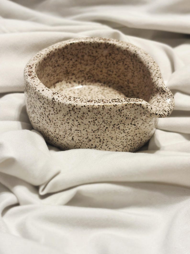 May include: A small, round ceramic bowl with a speckled beige and brown finish. The bowl features a distinct pouring spout on one side, a wide opening, and a smooth, curved exterior surface with a consistent pattern of dark brown flecks throughout.