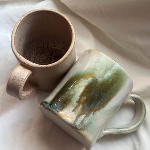 May include: Two ceramic mugs. One mug is upright, featuring a light beige speckled finish with a textured surface. The second mug is lying on its side, showcasing a glossy glaze with blended swirls of forest green, olive, and creamy white tones.