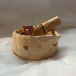May include: A ceramic matcha bowl with a brown bear figurine and two red hearts. The bowl is light beige with a textured surface. A bamboo matcha whisk is resting on the rim of the bowl.